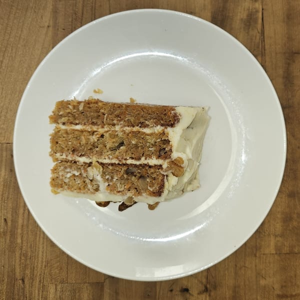 A slice of carrot cake with white frosting and walnuts on a white plate.