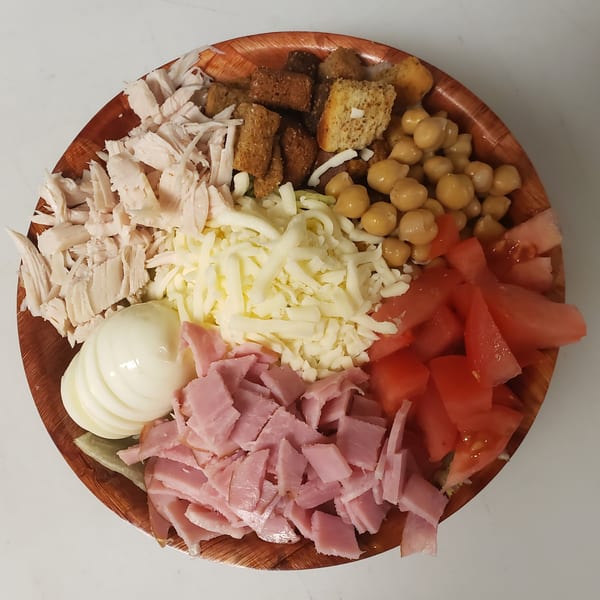 A wooden bowl filled with shredded chicken, chickpeas, croutons, cheese, tomatoes, ham, and hard-boiled egg.